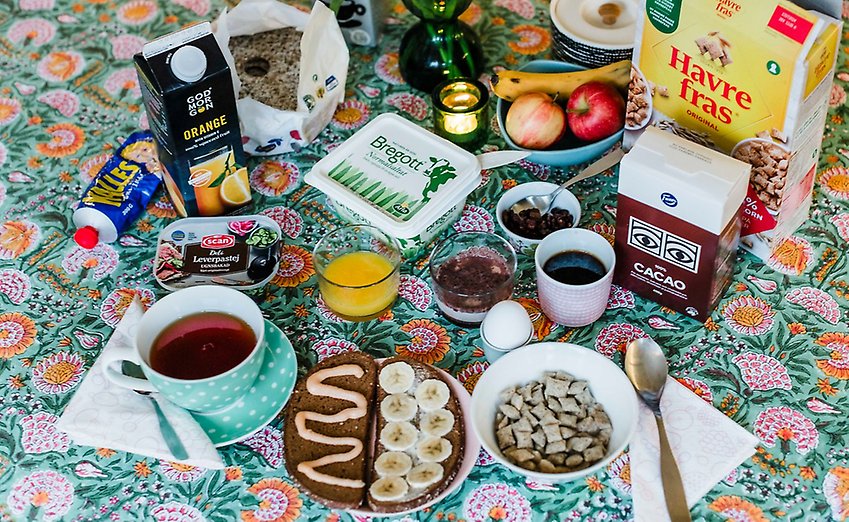 Ett bord med en blommig duk med en uppdukad frukost: kaffe, smör, yoghurt, flingor och annat.
