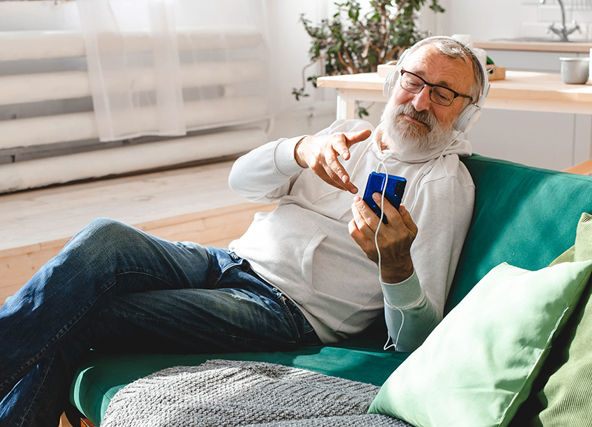 En äldre man med skägg sitter i en soffa med en mobiltelefon i handen.