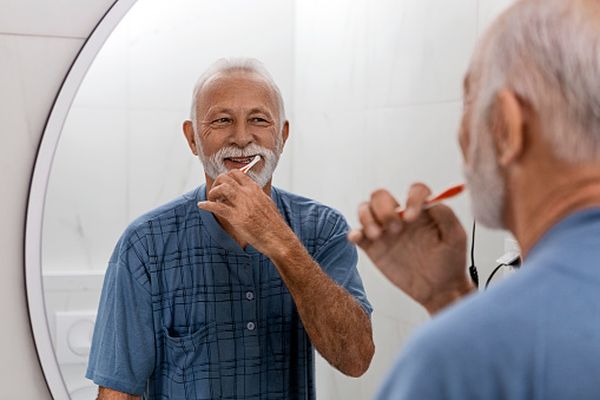 En man med vitt skägg står framför en spegel och borstar tänderna.