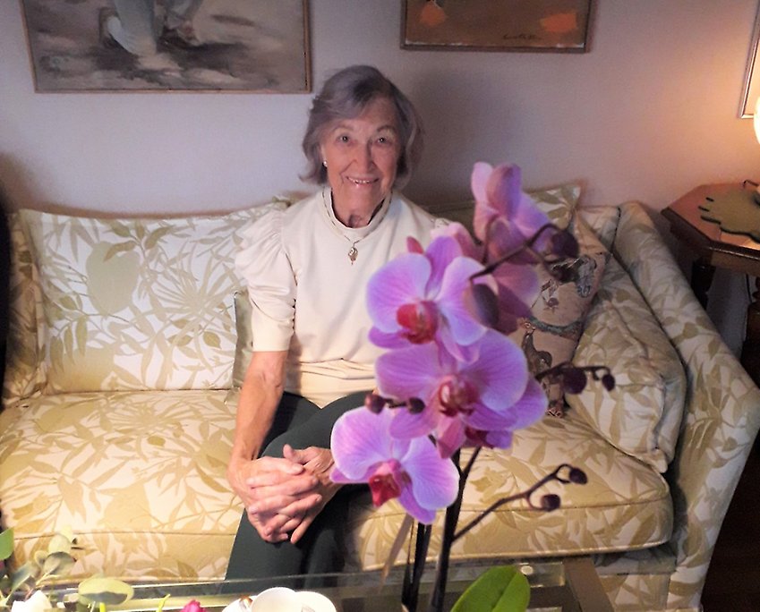 E kvinna sitter i en soffa. På bordet framför står en krukväxt - en orkidé.