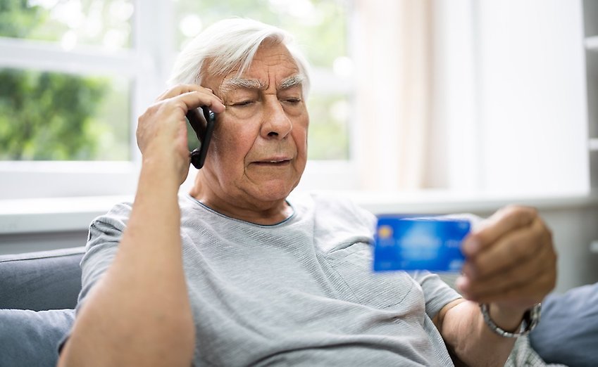 En äldre man sitter i en soffa och pratar i mobiltelefon, med ett bankkort i handen.