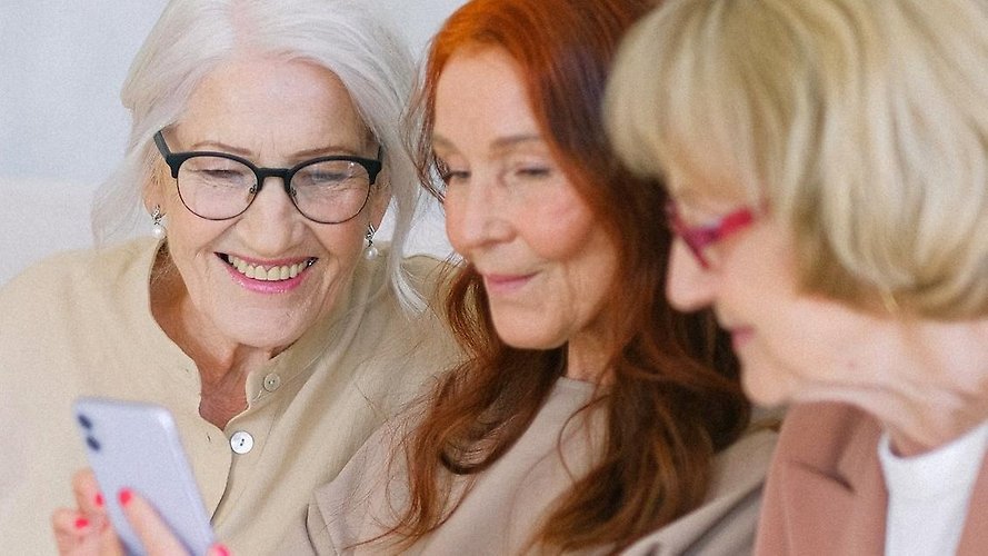Tre äldre kvinnor som tittar på en smart telefon, som den mittersta kvinnan håller i.