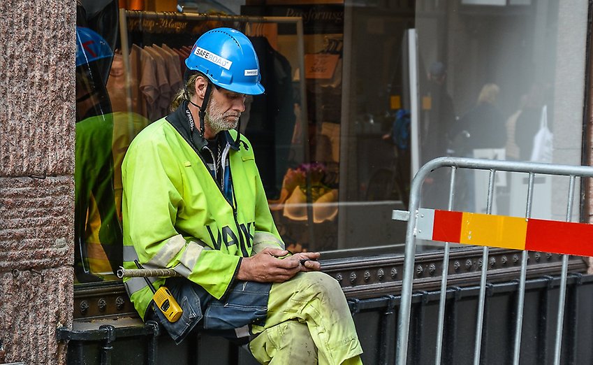 En byggnadsarbetare i gula arbetskläder sitter ute på gatan och tittar i sin mobiltelefon.