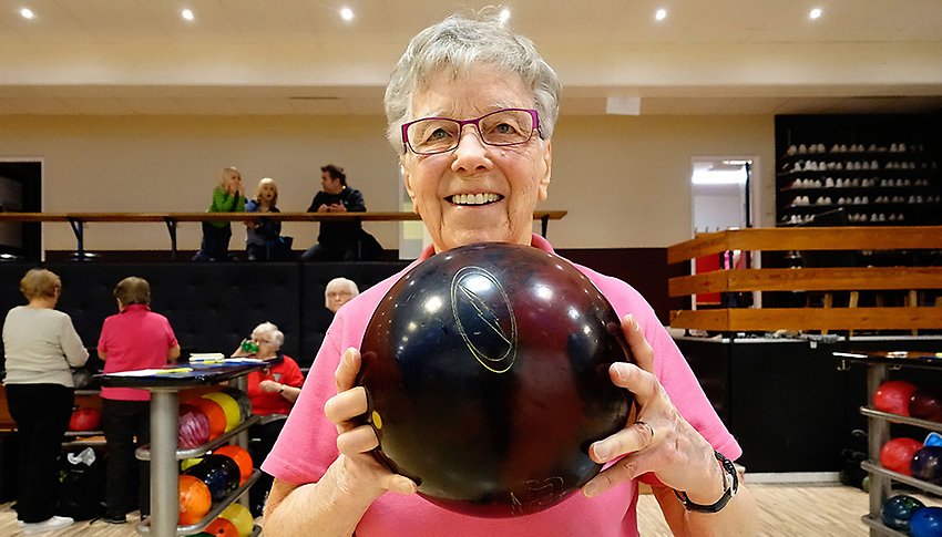 En äldre kvinnlig bowlingspelare står i en bowlinghall. Hon ler och håller ett bowlingklot i båda händerna.