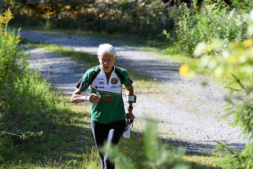 En äldre kvinna i orienteringskläder springer genom en solig lövskog.