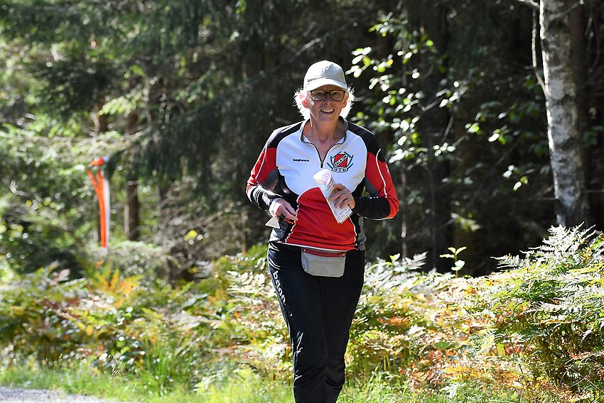 En äldre kvinna i orienteringskläder springer i en solig glänta i en lövskog.
