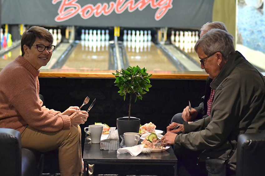 Två kvinnor sitter vid ett bord med smörgåsar och kaffekoppar, i bakgrunden ser man en bowlingbana.