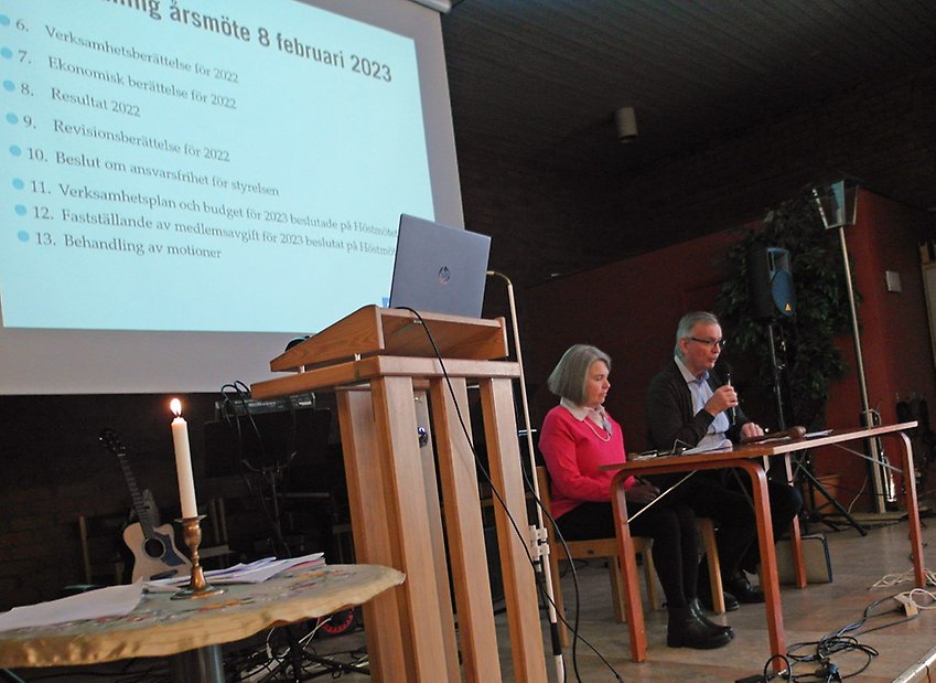Presidiet på PRO Tegs årsmöte, Tore Brännlund och Carola Johansson. De sitter vid ett bord framför på scenen medlemmarna med en storskärmsbild bakom som visar dagordning för årsmötet. Ett tänt ljus finns i förgrunden