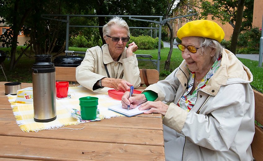Två äldre kvinnor sitter vid ett bord på en innergård och fika