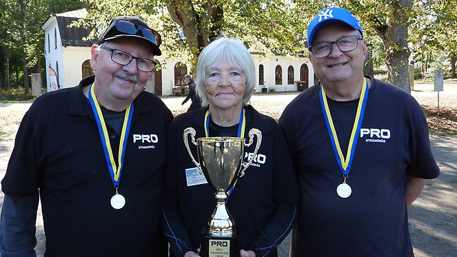 Tre äldre personer poserar med medaljer och en pokal