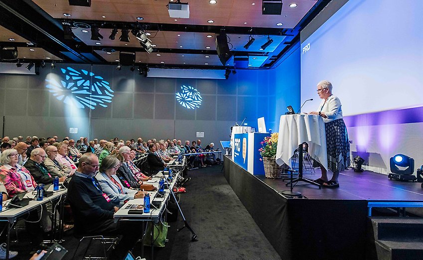 PRO:s ordförande Åsa Lindestam står i en talarstol på en inomhusscen med publik framför sig.