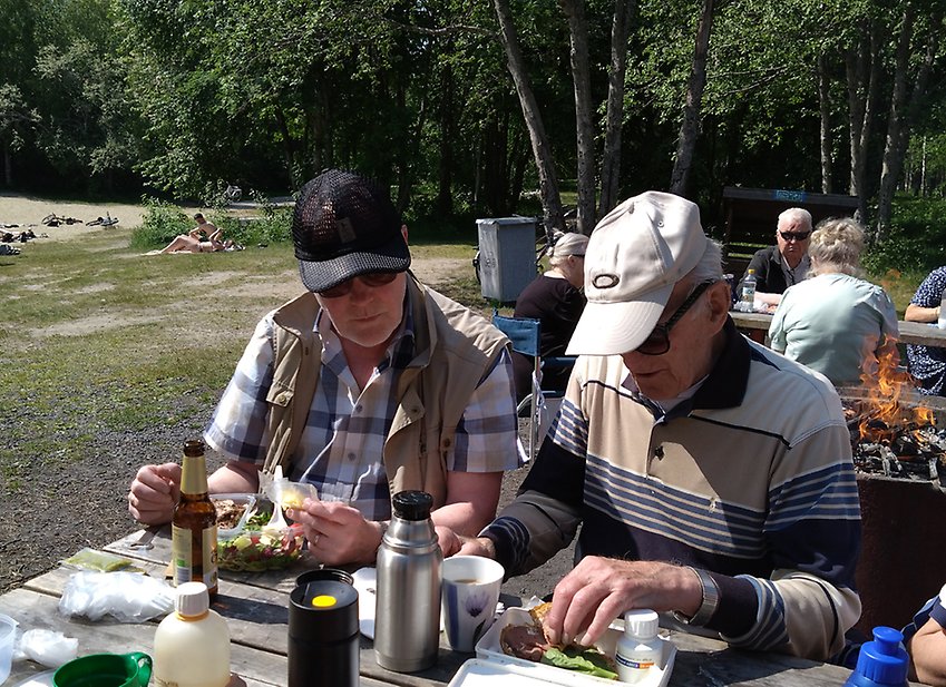 På PRO Tegs Picknick i det gröna samlades man runt borden för att avnjuta mat  och kaffe i trevligt sällskap. Man sitter vid bänkar som fanns uppställda vid skogsranden. utsikt mot badsjön
